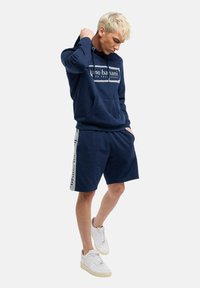Marineblauer Hoodie mit Fronttasche und Logo, kombiniert mit marineblauen Shorts mit seitlichen Akzenten und weißer Schrift. Weiße Sneaker runden den Look ab.