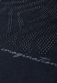 Tissu bleu marine avec des clous métalliques en relief formant des motifs abstraits et un texte manuscrit "imaginatioN" en police gris clair.