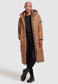 Abrigo tipo plumón largo y marrón con capucha, que presenta secciones acolchadas, una cremallera y botones a presión, llevado sobre pantalones negros y botines negros.