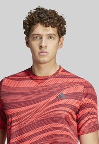 Camiseta atlética roja con un patrón de rayas onduladas, textura punteada y un logotipo oscuro en el lado izquierdo del pecho, mangas cortas y cuello redondo.