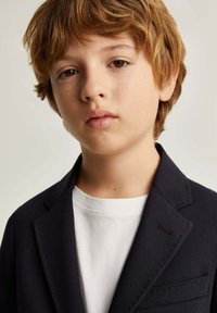 Blazer navy de tejido texturizado, con solapas con muesca y detalles en los bolsillos frontales sobre una camiseta blanca. Cabello corto y despeinado de color castaño claro.