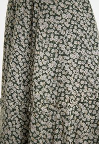 Groene stof met kleine, witte bloemmotieven en gouden accenten. Het ontwerp bevat plooien en een gerimpelde zoomafwerking.