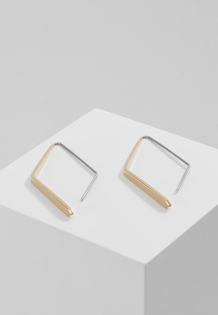 Paire de boucles d’oreilles géométriques avec une barre dorée et des crochets en métal argenté présentées sur un cube blanc sur fond gris.