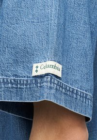 Μπλε τζιν ύφασμα με ραμμένη λευκή ετικέτα που δείχνει πράσινο κείμενο και λογότυπο που μοιάζει με "Columbia" σε μανίκι ενδύματος.