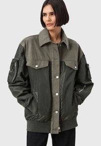 Giacca verde con una combinazione di finiture opache e lucide, ampie tasche frontali, chiusura a bottoni e una tasca laterale con zip sulla manica.