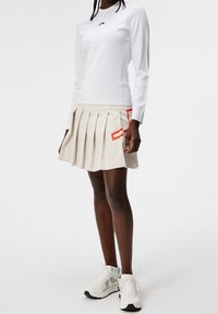 Mujer vestida con una blusa blanca de manga larga y una falda plisada beige con detalles rojos, combinada con zapatillas blancas gruesas, de pie sobre un fondo blanco.