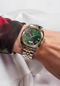 Reloj de oro y plata con una esfera verde, índices de números romanos y acentos de diamantes. Presenta un indicador de fecha y un bisel texturizado.