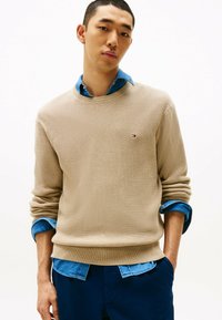 Beżowy sweter z fakturowanym splotem, okrągłym dekoltem i podwiniętymi mankietami, noszony na niebieskiej koszuli z denimu, z małym logo na piersi.