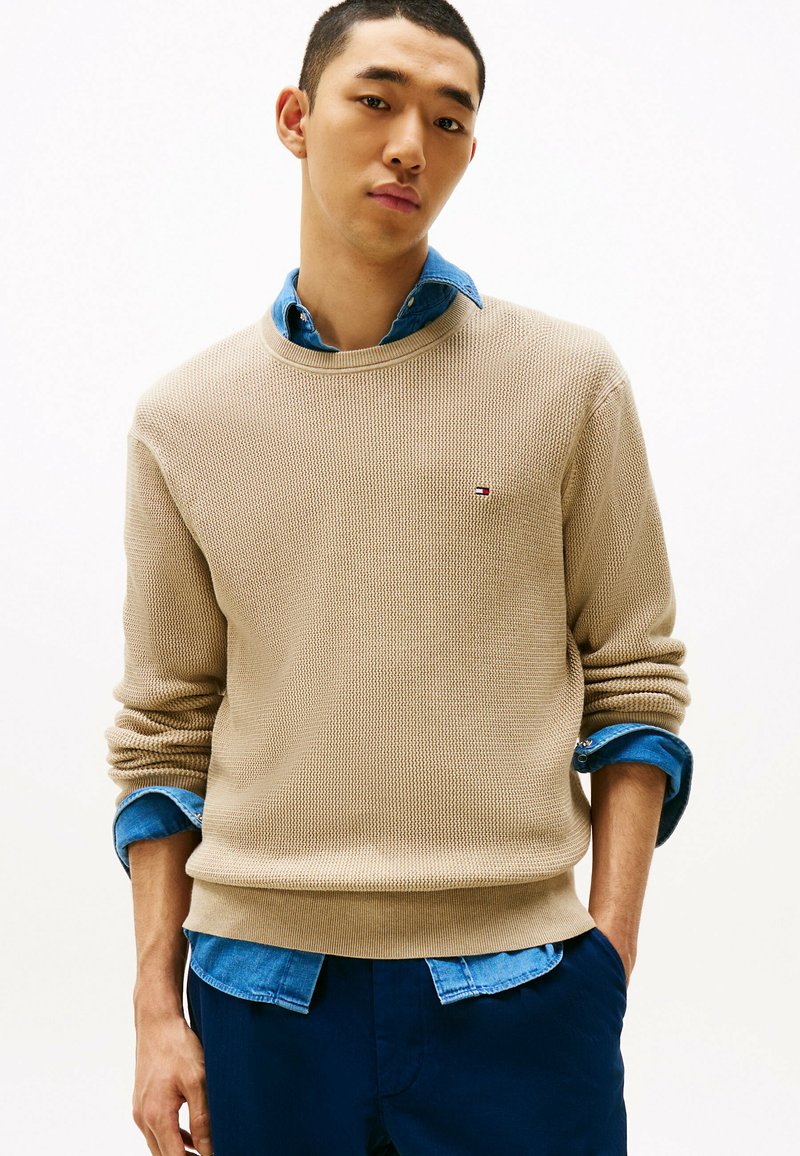 Beżowy sweter z fakturowanym splotem, okrągłym dekoltem i podwiniętymi mankietami, noszony na niebieskiej koszuli z denimu, z małym logo na piersi.