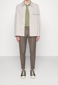 Světle béžová bunda bez límce s knoflíkovým zapínáním, zelené polo tričko, úzké hnědé kalhoty a černé tenisky s bílou podešví.