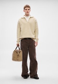 Béžový pletený sveter s golierom, hnedé nohavice širokého strihu a svetlohnedá kožená taška so zipsom a rúčkou, na jednoduchom pozadí.