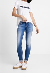 Mujer con una camiseta blanca de Hollister, jeans skinny azules deslavados arremangados en los puños y zapatillas de plataforma blancas contra un fondo sencillo.