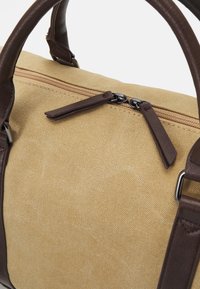 Canvas duffel bag in beige with dark brown leather accents. Features a zippered closure and padded handles for comfort. Textured surface.