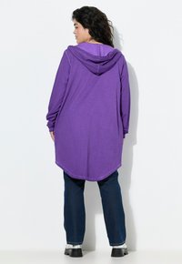 Lila Kapuzenpulli mit langen Ärmeln und abgerundetem Saum, kombiniert mit dunkelblauen Jeans und schwarzen Plateauschuhen. Der Stoff wirkt weich.