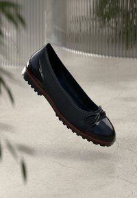 Black leather ballet flat with a glossy toe cap, textured body, and a decorative bow. Features a textured rubber sole for grip.
