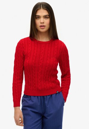 Roter gestrickter Pullover mit Zopf-Muster und langen Ärmeln. Rippstrick-Ausschnitt und -Manschetten. Kombiniert mit blauen gestreiften Hosen.
