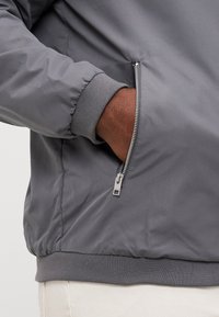 Chaqueta bomber gris con un bolsillo con cremallera, puños elásticos y un dobladillo acanalado, hecha de un material liso y ligero.