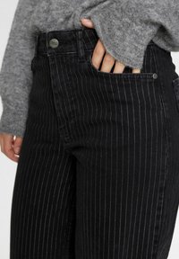 Svarta randiga jeans med hög midja, tillverkade av denim. Har en framficka med en silverknappdetalj, i kombination med en grå tröja.