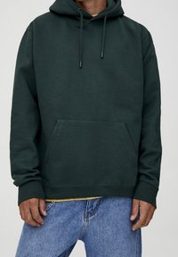 Personne portant un sweat à capuche vert foncé avec poche avant et un jean bleu, debout les mains détendues le long du corps.