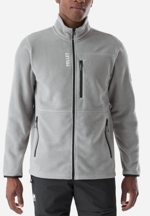 Man wearing light grey zip-up fleece jacket with black zipper pockets and black pants against white background.