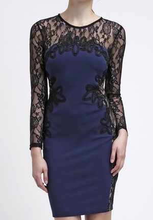 Femme portant une robe bleu marine ajustée avec des manches longues en dentelle noire et des détails en dentelle florale sur le corsage et les côtés.