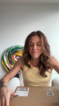 En kvinna i en gul linne sitter med en brun kartong på en vit stol med textur, händerna placerade som om hon håller på att öppna kartongen.