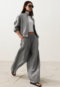 Femme portant un pantalon large gris à fines rayures et une veste assortie avec un crop top blanc, des lunettes de soleil noires et des tongs noires, marchant à l'intérieur.