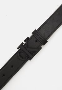 Čierny kožený opasok s veľkým matným čiernym logom "CK" ako sponou, s viditeľným šitím a otvormi na nastavenie. Textúra vyzerá hladko.