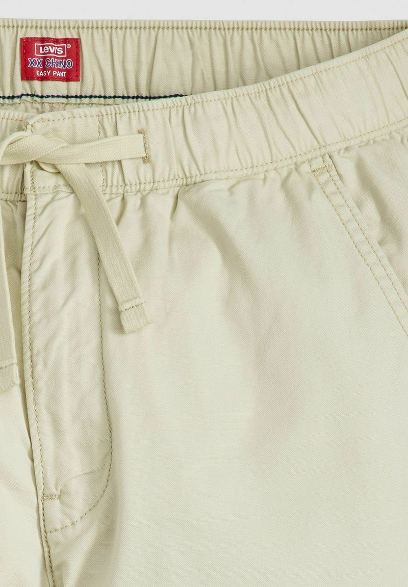 Chinos beige con cintura elástica y cordón, fabricados en una tela suave. Presentan una etiqueta roja con "Levi's XX Chino Easy Pant" y detalles de costura sutiles.