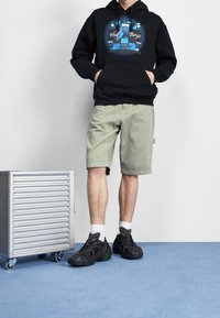 Sudadera negra con diseño gráfico azul, pantalones cortos verde claro y zapatillas negras escultóricas. El fondo incluye un carrito rodante plateado sobre un suelo azul.