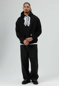 Zwarte hoodie met een groot wit sjaalontwerp aan de voorkant, voorzien van een voorzak en subtiele logo borduring. Zwarte wijde broek maakt het geheel compleet.