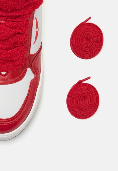 Red and white leather sneaker with textured red laces, rubber sole, and smooth toe box. Two additional rolled red laces positioned below.