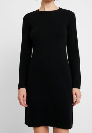 Vrouw draagt een effen zwart gebreide jurk met lange mouwen en een ronde hals, staand tegen een effen lichte achtergrond.