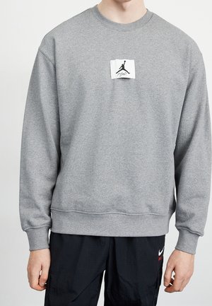 Person trägt einen grauen Pullover mit einem kleinen weißen Aufnäher mit schwarzem Jumpman-Logo und schwarze Hosen mit weißem Nike Air-Logo.