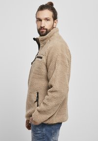 Beige fleecejakke med høj krave, struktureret overflade, sort lynlåslomme og sidelynlåse. Båret over et denimbukser.