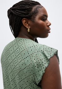 Jeune femme aux cheveux tressés portant des boucles d'oreilles créoles en or et un haut vert clair à manches courtes avec un motif ajouré, vue de profil.