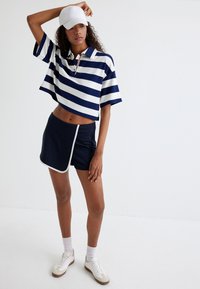 Mørkeblå og hvidstribet cropped polo skjorte, parret med mørkeblå shorts med hvid kant. Modellen bærer en hvid kasket og sneakers.