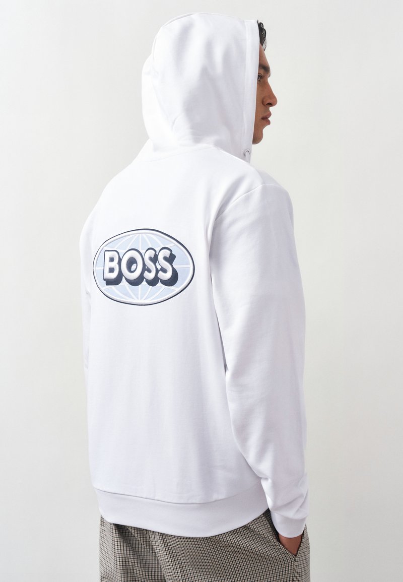 Homme portant un sweat à capuche blanc avec le logo "BOSS" dans le dos, debout de profil les mains dans les poches, fond uni.