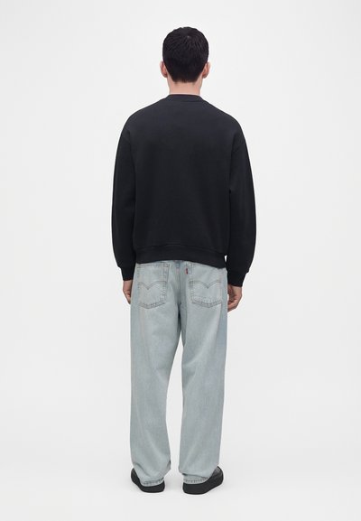 Homme debout, de dos, portant un sweat-shirt noir, un jean bleu clair ample et des chaussures noires sur un fond blanc uni.