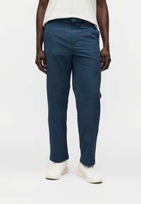 Παντελόνι σε σκούρο μπλε ναυτικό χρώμα από ελαφρύ ύφασμα, με κανονική γραμμή, θηλιές για ζώνη και πλαϊνές τσέπες. Φορεμένο με λευκά αθλητικά παπούτσια.