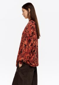 Przytulny, oversize sweter w czerwonobrązowym kolorze, wykonany z miękkiej, teksturowanej tkaniny z czarnym plamistym wzorem. Model ma na sobie ciemne spodnie.
