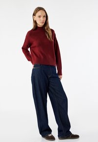 Burgundowa sweter dzianinowy z wysokim kołnierzem, zestawiony z luźnymi niebieskimi spodniami dżinsowymi z bocznymi kieszeniami i kontrastowym przeszyciem.