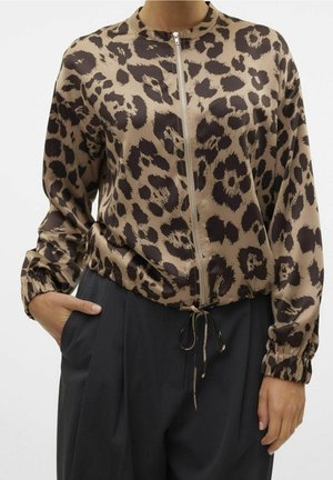 Vrouw draagt een bruin-zwart jaguarprint jack met ritssluiting en elastische manchetten, met zwarte broek, één hand in de zak en de andere ontspannen.