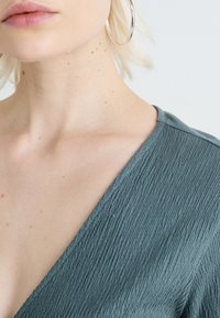 Texturerad, rynkad tyg i dämpad grön, som formar en v-ringning, tillsammans med stora, silverfärgade hoops örhängen mot en neutral bakgrund.