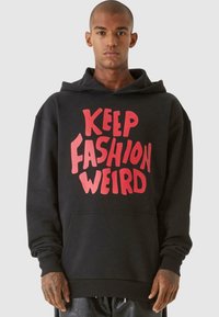 Felpa nera con una grande grafica rossa che recita "KEEP FASHION WEIRD." Presenta una tasca a marsupio e polsini a costine, realizzata in un spesso misto di tessuti.