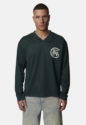 Man met kort haar draagt een donkergroen mesh sportshirt met lange mouwen en een wit logo op de borst en een lichtblauwe spijkerbroek.