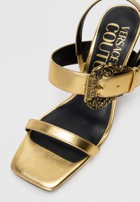Gold metallic heeled sandal with two thin straps and a square toe. Features a decorative buckle and black textured footbed.