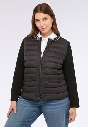 Donna con capelli castano chiaro che indossa una giacca a zip nera trapuntata con colletto borchiato sopra una camicia bianca e jeans blu, in posa su uno sfondo bianco.