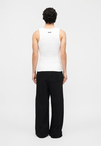 Personne aux cheveux bouclés portant un débardeur blanc moulant avec le texte "MSGM" dans le dos et un pantalon noir ample, debout devant un fond uni.