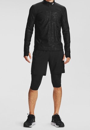Mann trägt schwarzen Sport-Langarm-Zip-Pullover, schwarze Shorts über schwarzen knielangen Leggings und schwarze Sportschuhe.